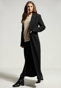 Cappotto lungo nero con bottoni, abbinato a un maglione beige. Il modello indossa pantaloni neri aderenti e stivaletti neri lucidi. Tessuto dalla consistenza liscia.