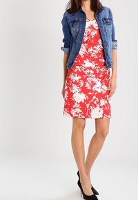 Czerwona sukienka w kwiaty z dekoltem w serek, zestawiona z niebieską dżinsową kurtką. Sukienka ma białe wzory kwiatowe i dopasowany krój. Czarne szpilki dopełniają całość.