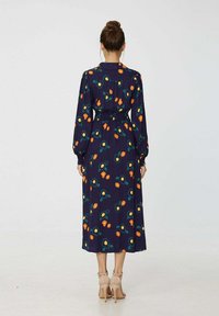 Robe midi florale navy à manches longues, taille smockée, avec des motifs floraux colorés orange et vert. Portée avec des sandales à talons beiges.