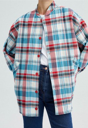 Personne portant une veste à carreaux ample avec des boutons rouges sur une chemise blanche et un jean bleu, les mains dans les poches, devant un fond uni.