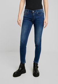 Jeans de mezclilla azul delgados con una cintura de altura media, bolsillos delanteros, detalles desgastados y una cremallera estándar, combinados con botas negras hasta el tobillo.
