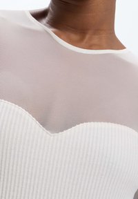 Haut beige clair avec un panneau supérieur en maille texturée et une section inférieure côtelée, ornée d'une couture ondulée, créant une apparence superposée.