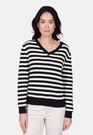 Femme debout portant un pull à col en V rayé noir et blanc et un pantalon de couleur claire sur un fond gris clair uni.