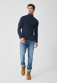 Tricou cu guler înalt de culoare bleumarin, croială slim, cu mâneci lungi; asortat cu blugi de culoare albastră deschisă și cizme de lucru negre cu șireturi. Design curat, textură netedă.