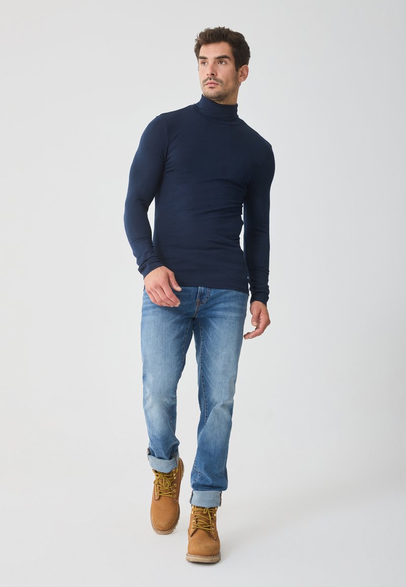 Tricou cu guler înalt de culoare bleumarin, croială slim, cu mâneci lungi; asortat cu blugi de culoare albastră deschisă și cizme de lucru negre cu șireturi. Design curat, textură netedă.