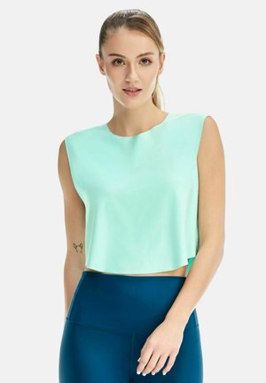 Junge Frau mit hellgrünem ärmellosem Crop-Top und dunkelblauen Leggings mit hoher Taille, stehend vor weißem Hintergrund.