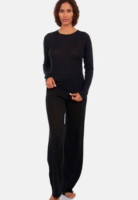 Femme portant un haut noir à manches longues et un pantalon noir ample, se tenant pieds nus devant un fond blanc uni.