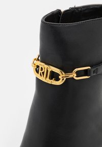Svart läder ankelboots med en guldkedja och "RL" logotypdetalj. Slät textur och strukturerad form.
