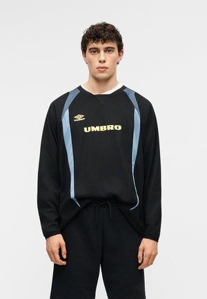 Νέος άνδρας με κοντά σγουρά σκούρα μαλλιά, φοράει μαύρο μακρυμάνικο πουκάμισο Umbro με γαλάζιες λεπτομέρειες και μαύρο παντελόνι, μπροστά σε απλό φόντο.
