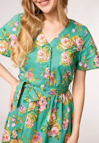 Grünes Floral-Kleid mit V-Ausschnitt, kurzen Ärmeln und gebundenem Bund. Floral-Muster in Pink und Gold verleihen dem weichen Stoff Details.