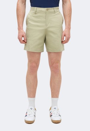 Person trägt beige knielange Shorts, weiße Sportsocken und weiße Sneakers mit marineblauen und roten Streifen, steht vor einem einfarbigen Hintergrund.