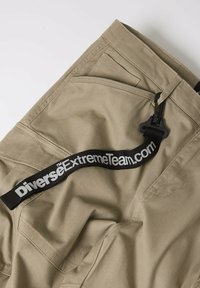 Khaki szorty cargo z dużymi kieszeniami, gładką teksturą i czarnym paskiem z napisem "DiverseExtremeTeam.com" w białych literach.