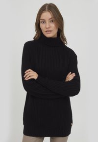 Pull à col montant noir avec un design en maille texturée et épaisse, et des bords ribbed au niveau des poignets et de l'ourlet. Présente un petit logo sur le côté inférieur.