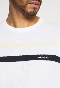 Biały sweter z dzianiny z poziomymi paskami w kolorze żółtym i granatowym, z logo "JACK&JONES" w żółtym kolorze na przodzie. Okrągły dekolt.