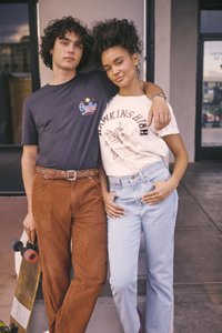 Két személy áll egymás mellett. A bal oldalon álló személy sötét, mintás pólót és barna kordbársony nadrágot visel; kezében gördeszkát tart. A jobb oldalon álló személy krémszínű pólót fekete felirattal és világoskék farmert visel.