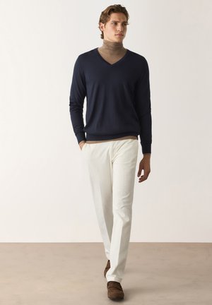 Jeune homme marchant portant un pull en V bleu marine sur un col roulé marron, pantalon blanc et mocassins marron sur un fond uni.
