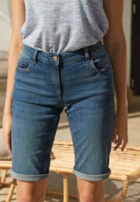 Personne portant un short en jean retroussé et un haut sans manches gris clair, debout près d'un banc en bois extérieur.