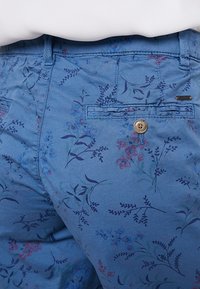 Pantalon en coton bleu clair avec un motif floral en nuances de rose et de violet, doté d'une poche arrière avec une fermeture à bouton rond.