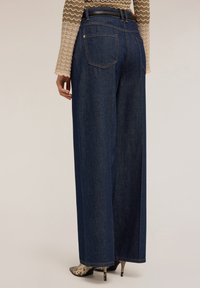 Pantalones vaqueros de mezclilla de pierna ancha en un tono azul oscuro con cintura alta, que presentan bolsillos traseros de parche y una textura suave. Combinados con tacones con estampado de serpiente.