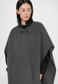 Sivi vuneni ogrtač s visokim ovratnikom, s crnim kopčanjem. Tkanina je glatke teksture, nošena preko crnog džempera s teksturom.