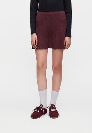 Personne portant une jupe en maille torsadée bordeaux, un haut noir à manches longues, des chaussettes blanches et des baskets bordeaux avec des lacets blancs.
