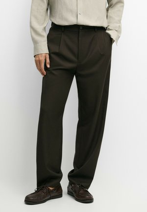 Homme portant un pantalon plissé marron foncé, une chemise beige clair rentrée, et des chaussures en cuir marron foncé lacées, debout les mains dans les poches.