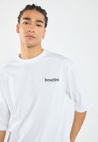 Hvit bomulls t-skjorte med rund hals og korte ermer, prydet med svart og grønn "houdini" logo på venstre bryst.