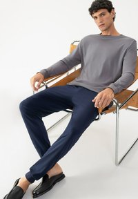 Grijze lange mouwen top, marineblauwe broek met een gladde textuur en zwarte instappers. Model zit op een gebogen houten stoel met chroom benen.