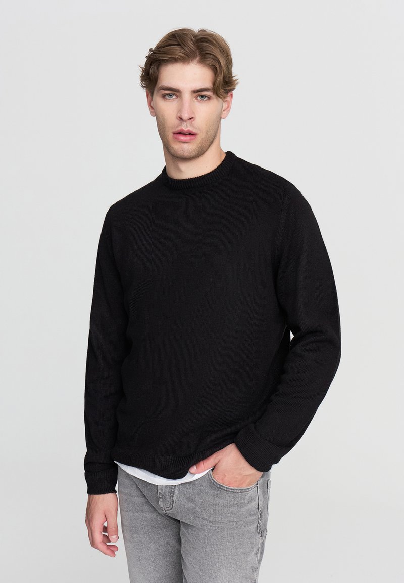 Maglione nero in maglia con scollatura a girocollo, a costine nei polsini e nell'orlo. Indossato sopra una camicia bianca, abbinato a jeans grigio chiaro. Texture liscia.