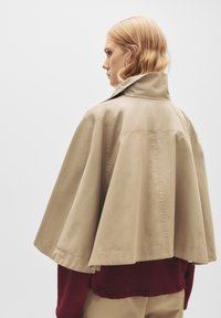 Cappotto color beige con un colletto alto, dotato di maniche ampie e una texture liscia. Indossato sopra un indumento di un profondo color bordeaux. Design minimalista.