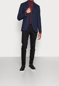 Saco a cuadros en tono azul marino con dos botones, combinado con un cuello alto color burdeos, pantalones ajustados negros y zapatillas negras con suelas blancas.