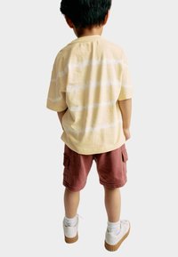 Camiseta amarilla a rayas con mangas cortas, combinada con pantalones cortos cargo rojos. Zapatillas blancas con suela de goma, calcetines blancos cortos.