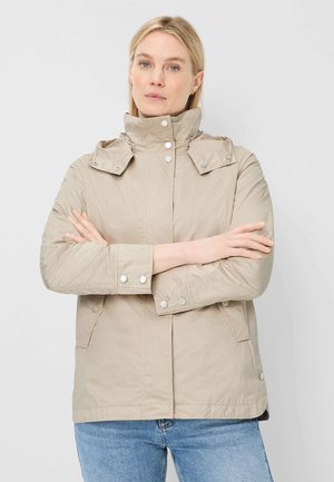 Frau mit verschränkten Armen, die eine beige Jacke mit hohem Kragen und blaue Jeans trägt, steht vor einem schlichten hellen Hintergrund.