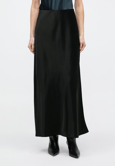 Kobieta ubrana w długą czarną satynową spódnicę, do której dopasowane są czarne botki z zaostrzonym noskiem, na jednolitym białym tle.