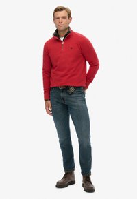 Rode fleece pullover met een kwart rits, gelaagd over een zwart-groene geruite shirt, gecombineerd met blauwe jeans en bruine veterschoenen.