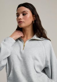 Grå zip-tröja med gulddragkedja, featuring en framficka och en avslappnad passform. Mjukt tyg med synlig sömdetalj.