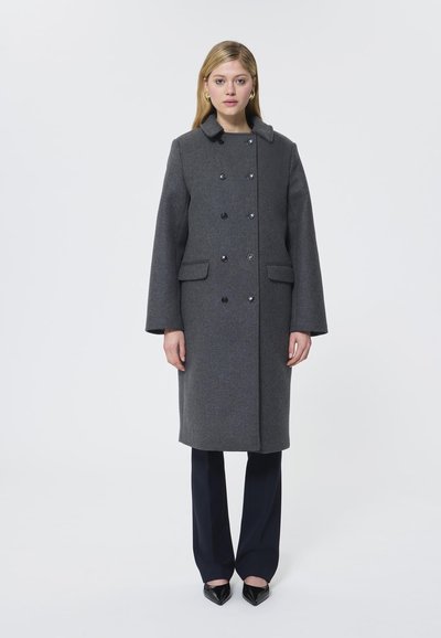 Cappotto in lana grigia con colletto, bottoni doppi, due tasche frontali e silhouette dritta. Abbinato a pantaloni scuri e scarpe a punta.