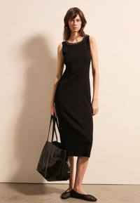 Abito senza maniche in maglia nera con scollatura decorata da perline, abbinato a una borsa a tracolla nera lucida e a scarpe slip-on in tessuto intrecciato nere. Texture liscia.