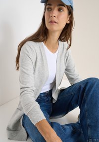 Cardigan grigio chiaro a maglia con apertura frontale e polsini a coste, abbinato a una t-shirt bianca e jeans in denim blu. Sfondo neutro.