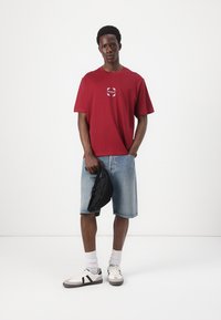 Red short-sleeve t-shirt featuring a white circular logo, paired with blue denim shorts and black crossbody bag. Gray sneakers with black accents.