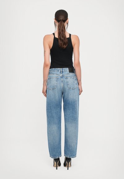 Calças de ganga de última subida em azul claro, com um corte descontraído, cinco bolsos e costura standard. Usadas com uma parte de cima preta sem mangas.