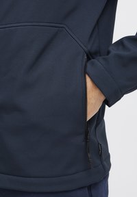 Marineblå softshell-jakke med glidelåslomme, glatt tekstur og en tettsittende design med en hemmet finish.