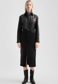 Vrouw in een zwart leren knopenoverhemd, zwarte midi-rok met een opening aan de voorkant, zwarte bandana en zwarte laarzen tot halverwege de kuit tegen een witte achtergrond.