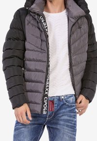Chaqueta acolchada con un diseño a bloques de color en secciones negra y gris, cierre de cremallera y detalles de marca a lo largo del frente y la capucha.
