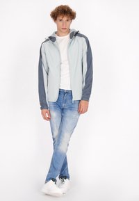 Giacca zip blu chiaro con maniche blu scuro, jeans slim-fit e sneakers bianche. Il tessuto appare liscio con una finitura opaca.