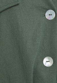 Tissu vert à finition texturée avec un grand bouton de couleur claire et un col. Le matériau semble durable et tissé.