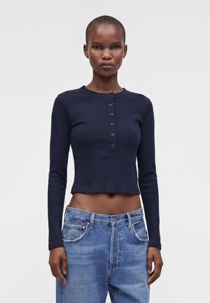 Jeune femme aux cheveux courts portant un haut court bleu marine à manches longues et boutonné ainsi qu'un jean bleu taille haute sur un fond uni.
