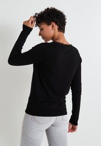 Pull noir à manches longues avec un col festonné, des poignets côtelés et une texture douce, associé à un jean gris clair.