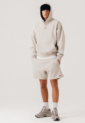 Joven con sudadera beige con capucha puesta, pantalones cortos a juego, calcetines blancos, zapatillas grises y gorro negro, de pie con las manos en los bolsillos.
