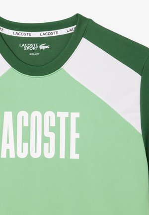 Maglia sportiva verde con collo a giro, pannelli delle spalle bianchi a contrasto e grande scritta "LACOSTE" in bianco. Design a vestibilità regolare.